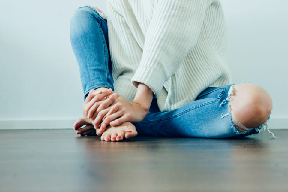 Headless woman sits on wooden floor with ankles crossed and hands on ankles working on PMS