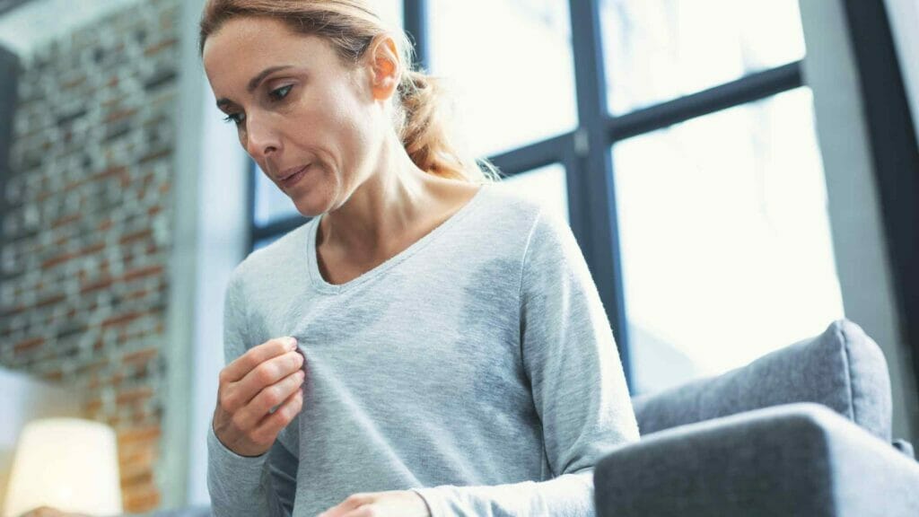 A woman in a grey top with sweat stains sitting and cooling herself by shaking her top to help ease the perimenopause or menopause symptoms