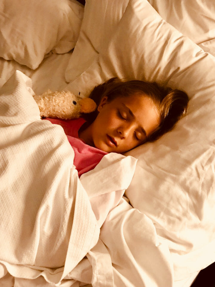 IMage of a young girl asleep in bed with a fluffy duck toy, sleeping soundly after her parents followed the tips to help your kids sleep better