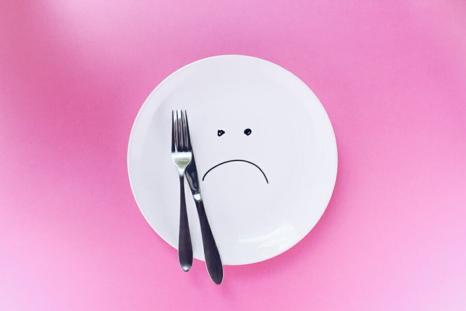 Image of an empty plate with a knife and fork and a frowning face to indicate that skipping meals is not helpful in helping menstrual cramps.