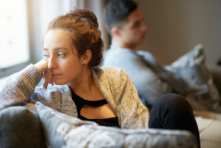 Woman looks out window due to relationship stress with her partner in the background also looking stressed