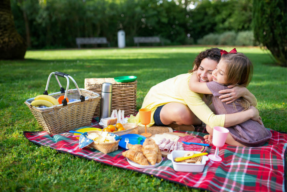 Image of a mother hugging her daughter whilst at a picnic smiling showing the benefits of hugging your child