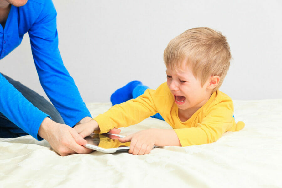 Image of a young white boy with blonde hair in a yellow long sleeve top and blue pants laying on a bed struggling with a headless parent in a blue long sleeve shirt over a tablet whilst the parent is likely wonder why your child acts out