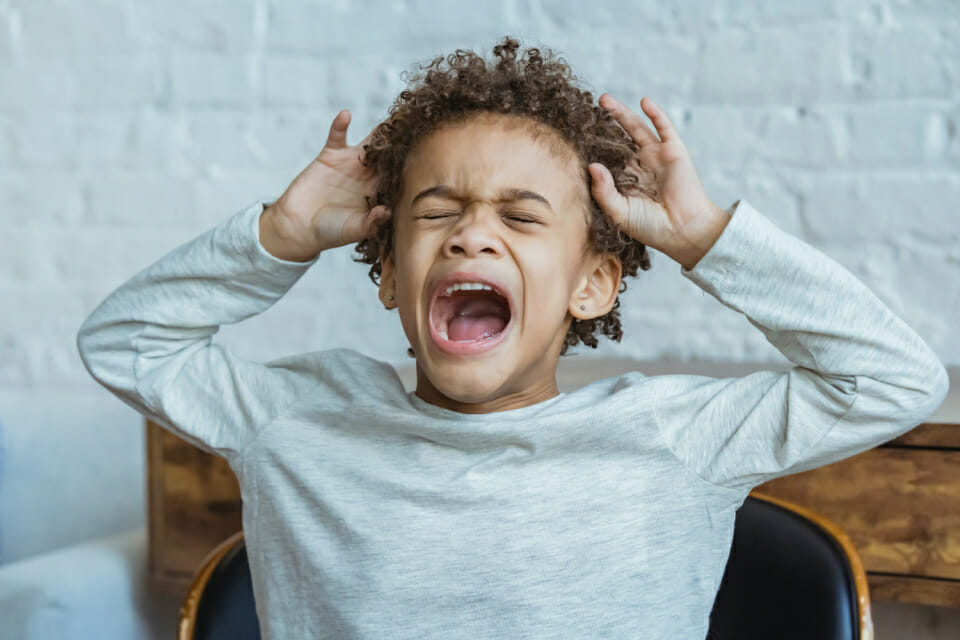 Image of a mixed race boy of colour screaming with a white brick wall behind probably triggering the question of why do kids act out