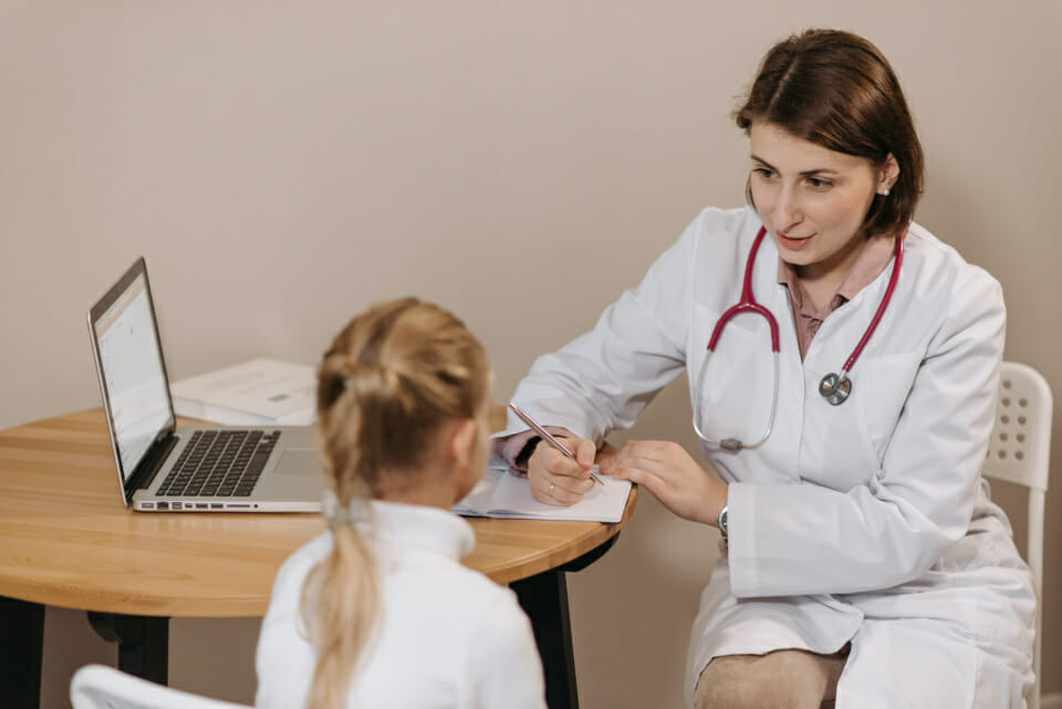 Image of a white female doctor with brown shoulder length hair wearing a white lab coat and stethoscope sitting at a table that has a laptop on it and writing with pen and paper with a young white blonde haired girl in a white shirt who's back is to the camera likely discussing why do kids act out