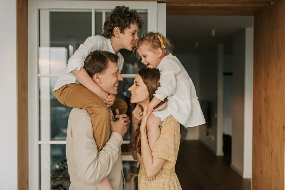 Image of a white mother with her young daughter on her shoulders and a white father with his young son on his shoulders sharing positivity to bring the family closer together.