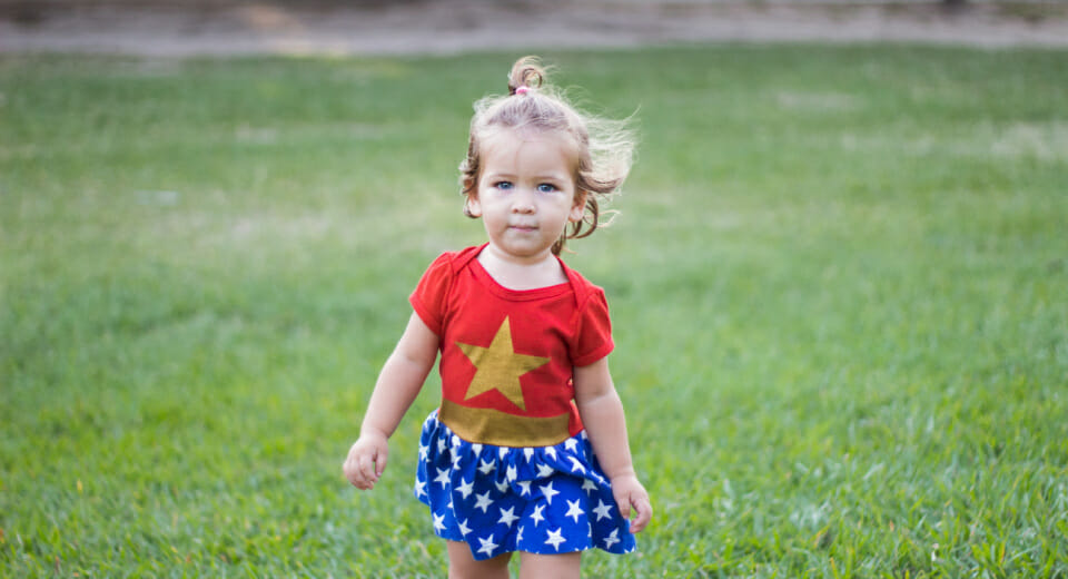 Little white girl wearing a wonder woman dress walking on a grass field looking straight at the camera because you improve your kid's self-awareness