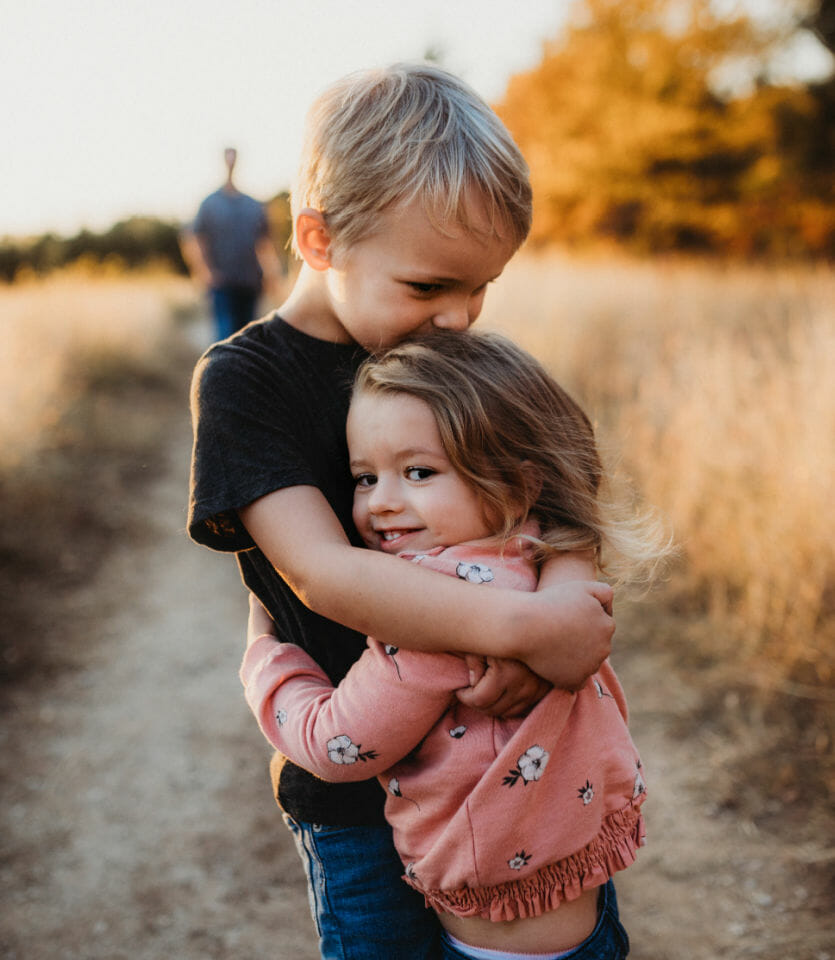 A young boy and girl hug in a field in the foreground with an adult walking behind giving them the space to improve your child's self-awareness