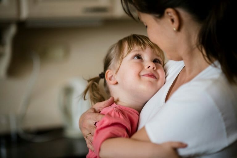 Image of a white mother hugging her young daughter who is smiling up at you showcasing the benefits of hugging your child